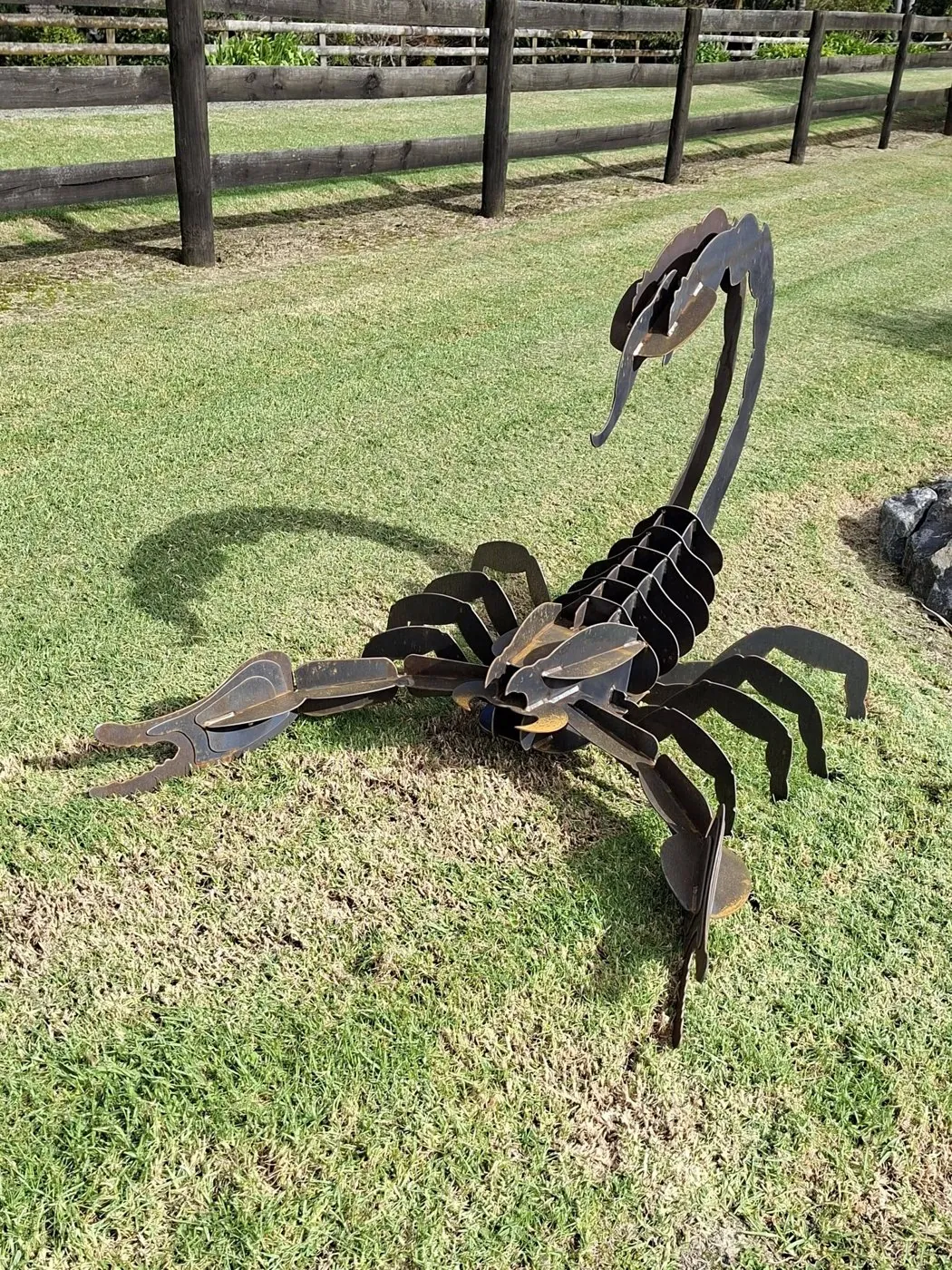 Metal scorpion sculpture on grass beside a timber fence.