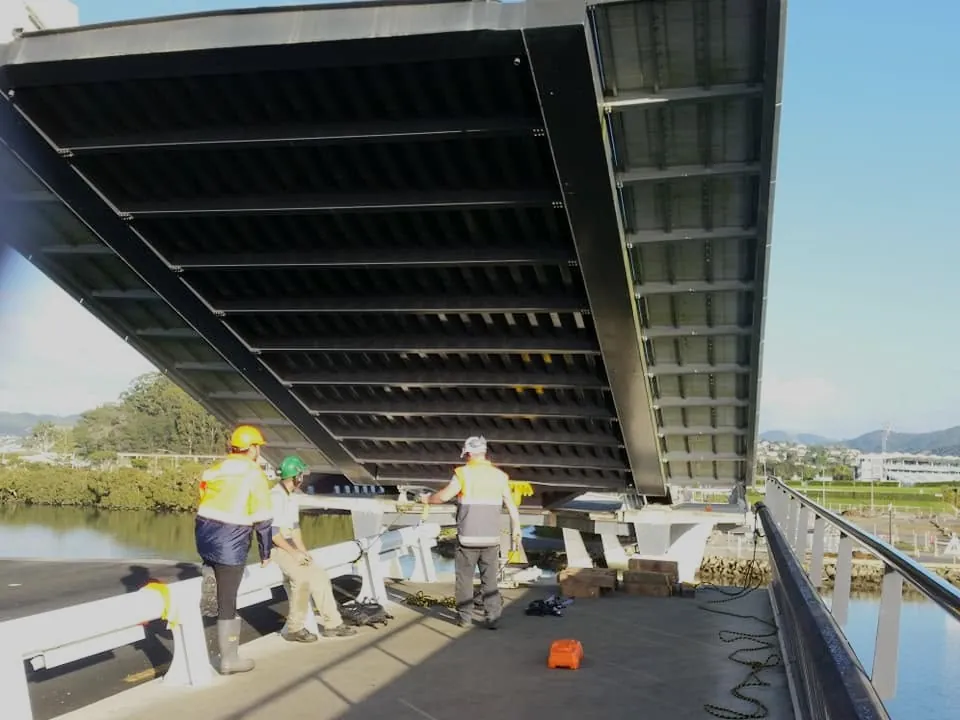 Workers and equipment beneath a bridge beside a river.